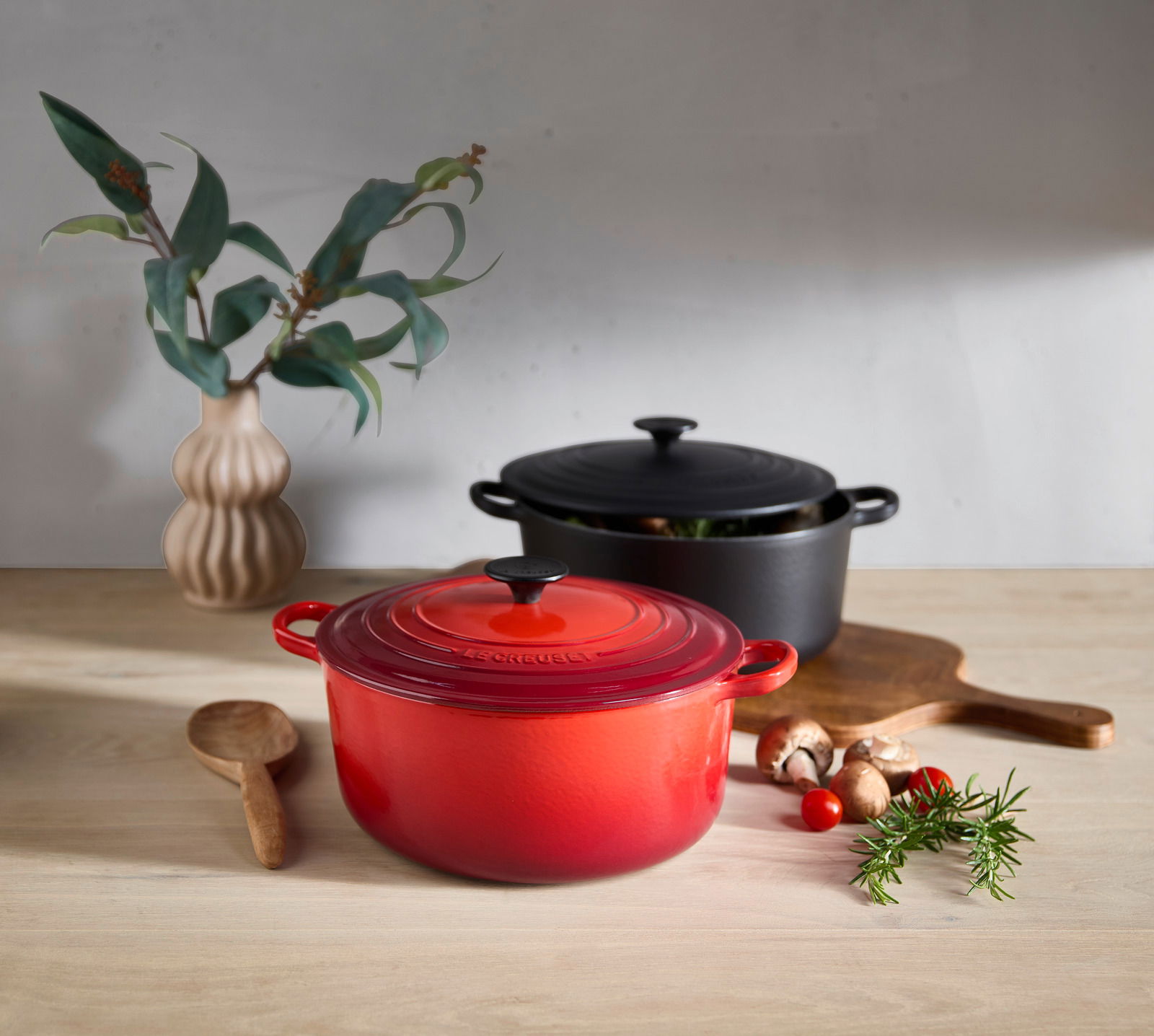 LE CREUSET Bräter 26cm Kirschrot TRADITION Roter Bräter mit Deckel in Kirschrot, auf einem Holztisch, umgeben von frischen Zutaten und einem Holzlöffel, aus einer leicht seitlichen Perspektive fotografiert.