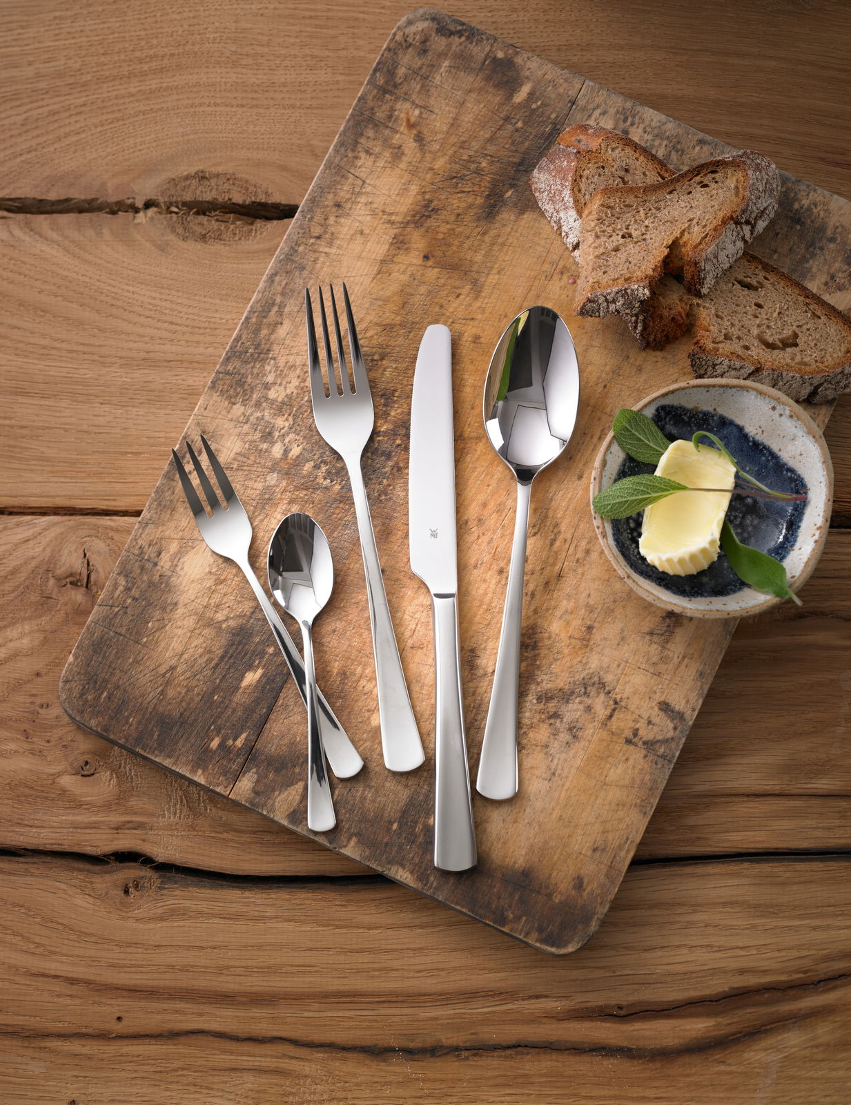 Besteckset Denver 60-tlg. auf einem rustikalen Holzbrett, bestehend aus Messer, Gabel, Löffel und Teelöffel, mit Brotscheiben und Butter im Hintergrund, aus der Vogelperspektive fotografiert.