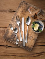 Besteckset Denver 60-tlg. auf einem rustikalen Holzbrett, bestehend aus Messer, Gabel, Löffel und Teelöffel, mit Brotscheiben und Butter im Hintergrund, aus der Vogelperspektive fotografiert.