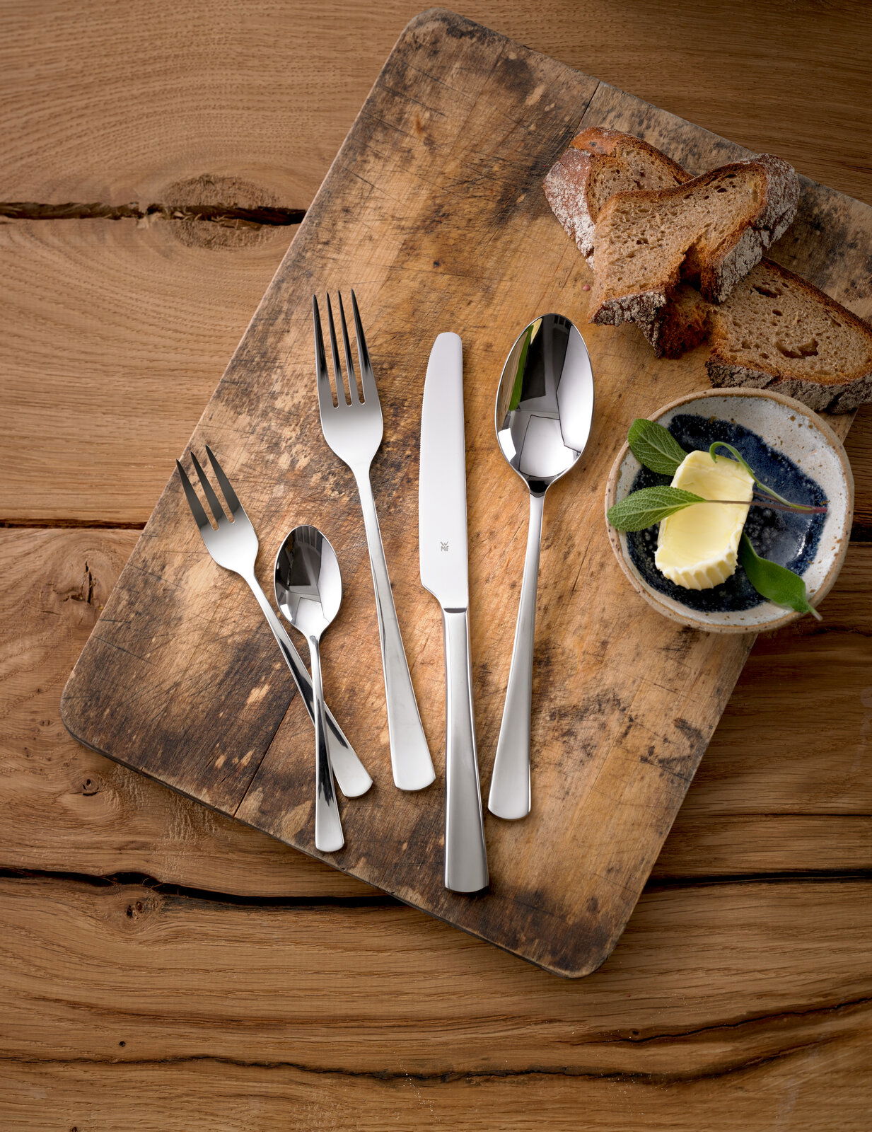 Besteckset Denver 60-tlg. auf einem rustikalen Holzbrett, bestehend aus Messer, Gabel, Löffel und Teelöffel, mit Brotscheiben und Butter im Hintergrund, aus der Vogelperspektive fotografiert.