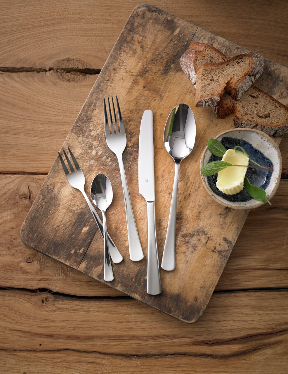 Besteckset Denver 60-tlg. auf einem rustikalen Holzbrett, bestehend aus Messer, Gabel, Löffel und Teelöffel, mit Brotscheiben und Butter im Hintergrund, aus der Vogelperspektive fotografiert.