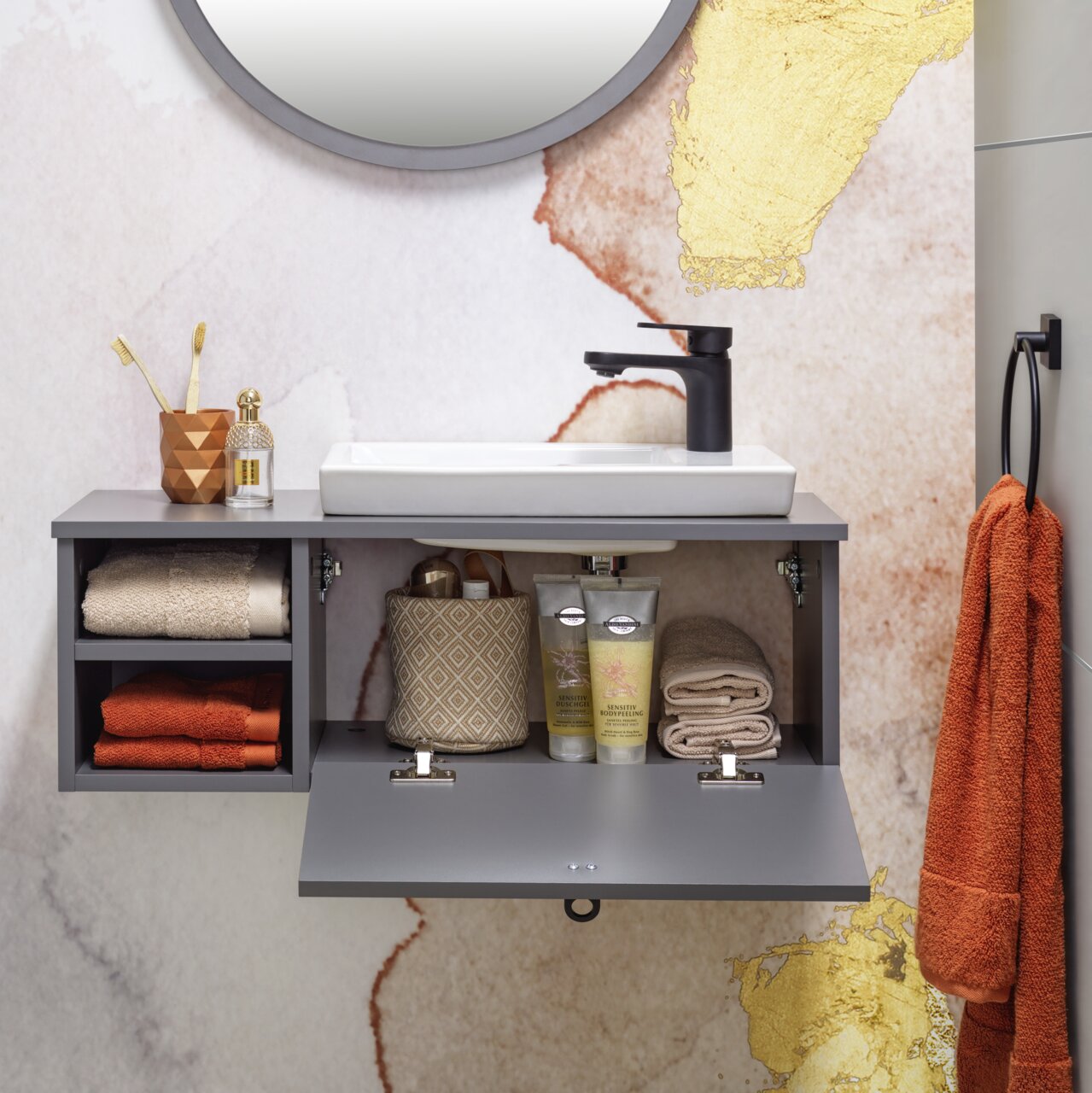 Modernes Waschtisch-Set mit grauem Schrank, weißem Waschbecken und schwarzem Wasserhahn, frontal fotografiert. Dekoriert mit Handtüchern, Zahnbürsten und Pflegeprodukten.