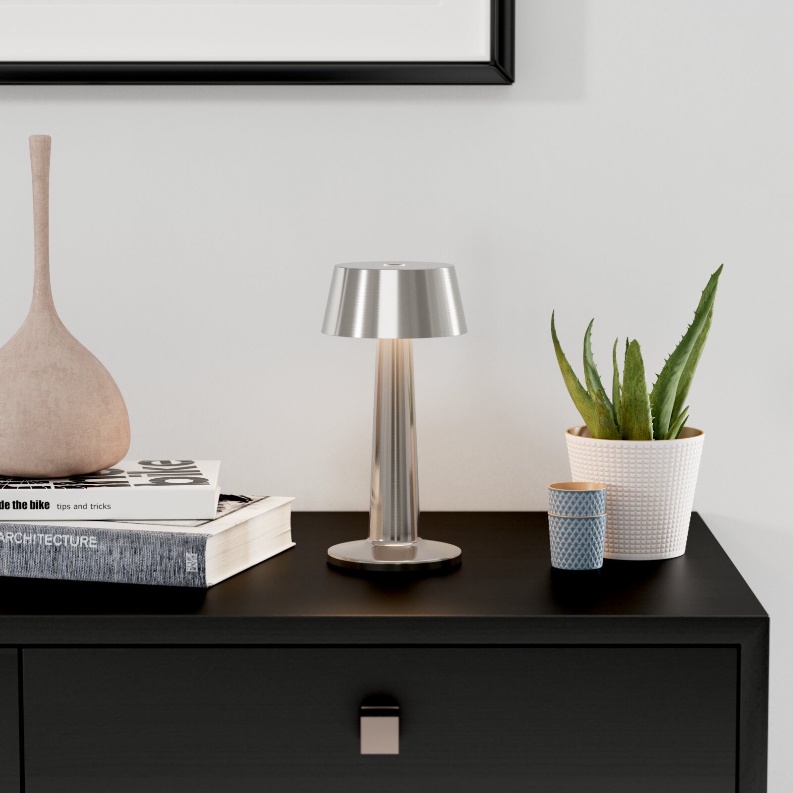 Moderne silberne Tischleuchte aus Metall mit konischem Schirm auf schwarzem Sideboard, frontale Perspektive; daneben Bücherstapel, Dekovase und Topfpflanze vor heller Wand