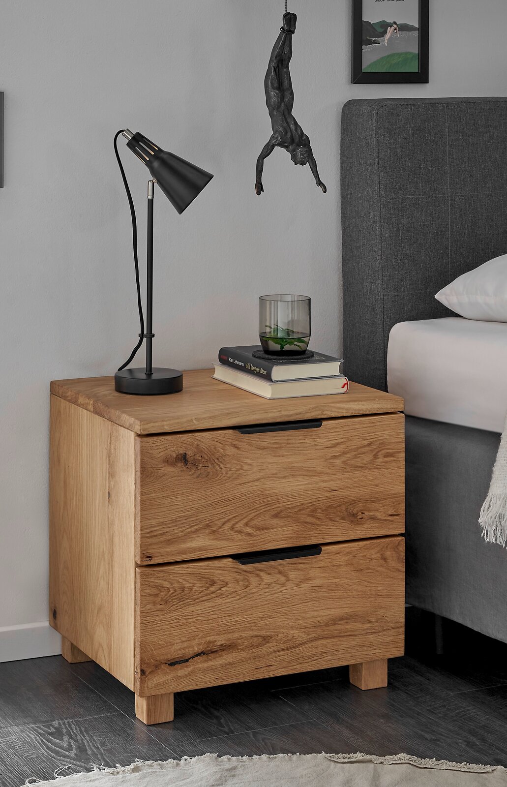 Nachtkommode aus Holz mit zwei Schubladen, darauf eine schwarze Lampe, Bücher und ein Glas. Perspektive von vorne links.