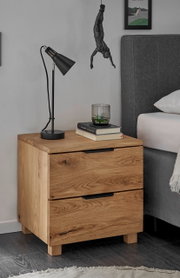 Nachtkommode aus Holz mit zwei Schubladen, darauf eine schwarze Lampe, Bücher und ein Glas. Perspektive von vorne links.