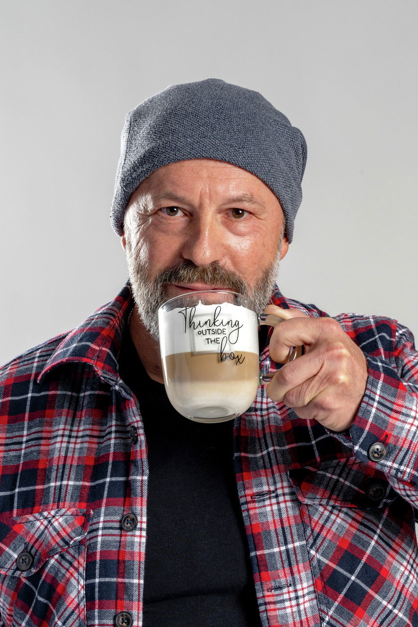 Mann mit grauer Mütze und kariertem Hemd hält eine Tasse mit der Aufschrift 'Thinking outside the box' in der Hand, frontal aufgenommen.