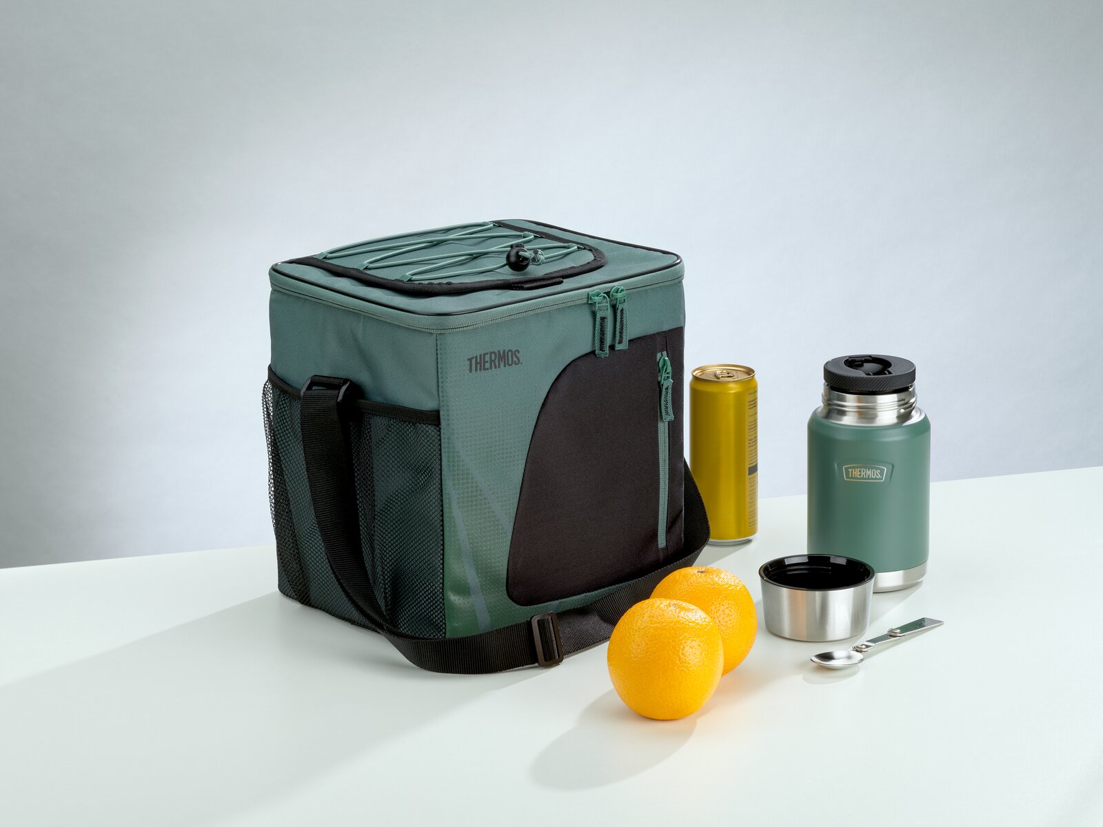 Grüne THERMOS Kühltasche mit Schultergurt und Reißverschlussfächern, aus schräger Vorderansicht neben Isolierbehälter, Getränkedose, zwei Orangen, Becher und Löffel präsentiert