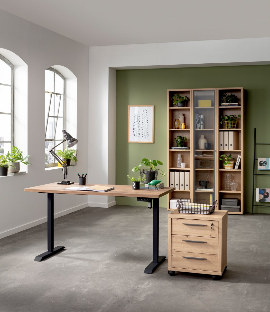 Moderner höhenverstellbarer Schreibtisch mit schwarzem elektrischen Fußgestell und Holzplatte in einem hellen Büro mit Pflanzen und Regalen, seitliche Perspektive.