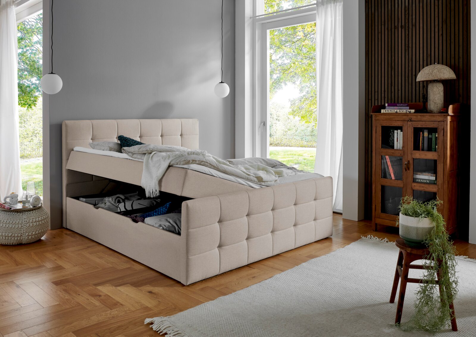 Een beige boxbed met opbergruimte, in geopende toestand, in een moderne slaapkamer met houten vloer en grote ramen, gezien vanaf de zijkant.