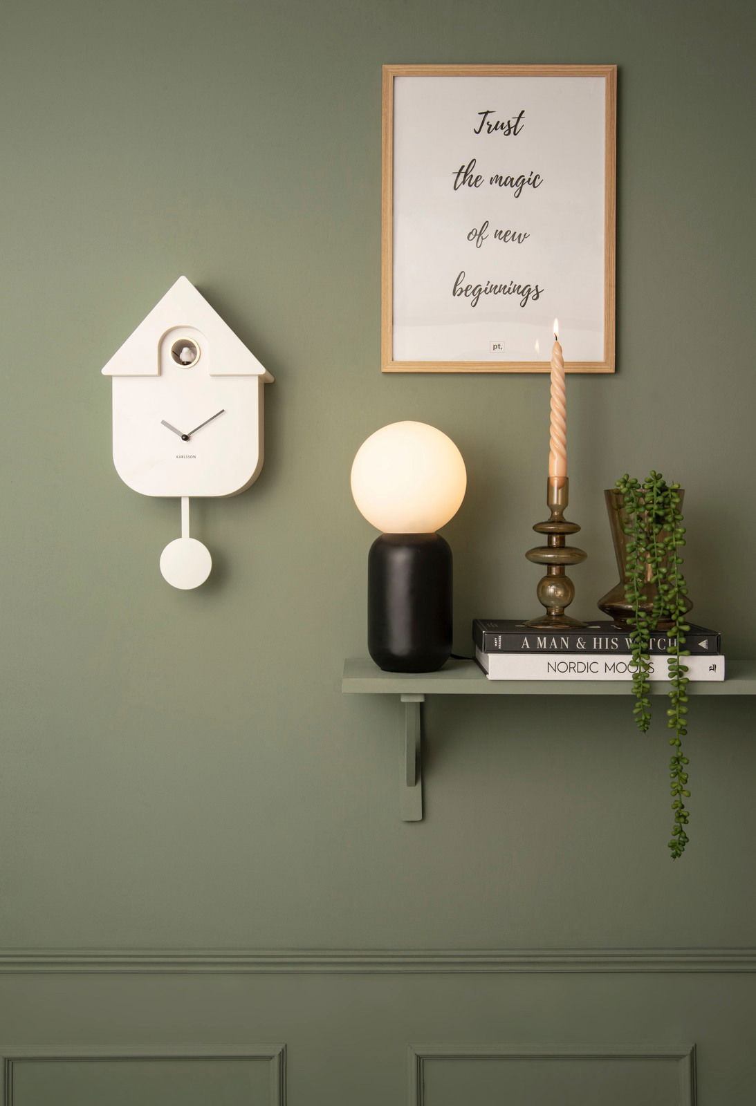 KARLSSON Wandklok Moderne Wanduhr in Form eines Hauses mit Pendel, an einer grünen Wand montiert, neben einem Regal mit Lampe, Kerze und Pflanzen, aus frontaler Perspektive.