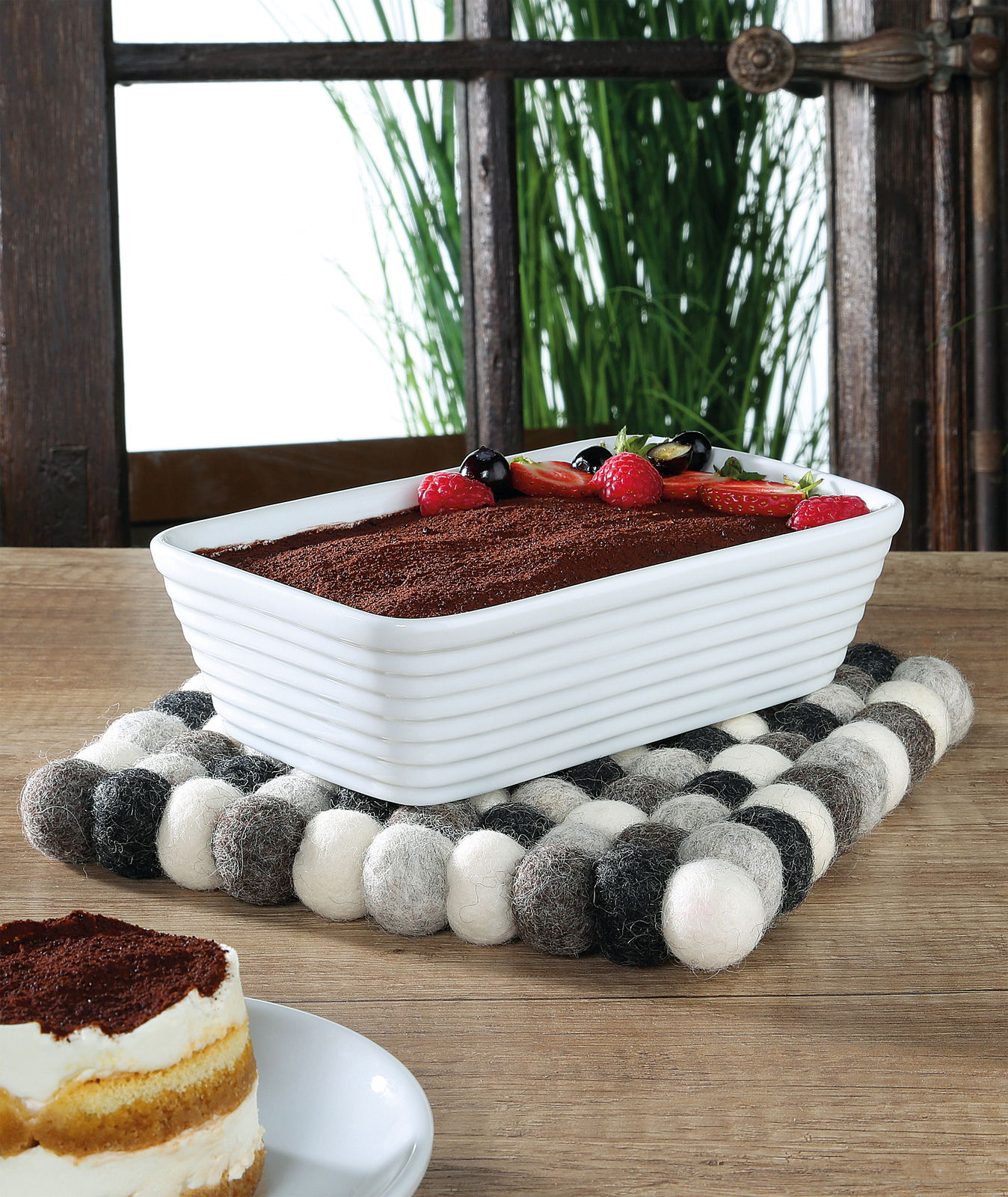 Quadratischer Untersetzer Lana Grigio aus Filzkugeln in Grautönen, auf einem Holztisch mit einer weißen Auflaufform und einem Stück Tiramisu, seitliche Perspektive.
