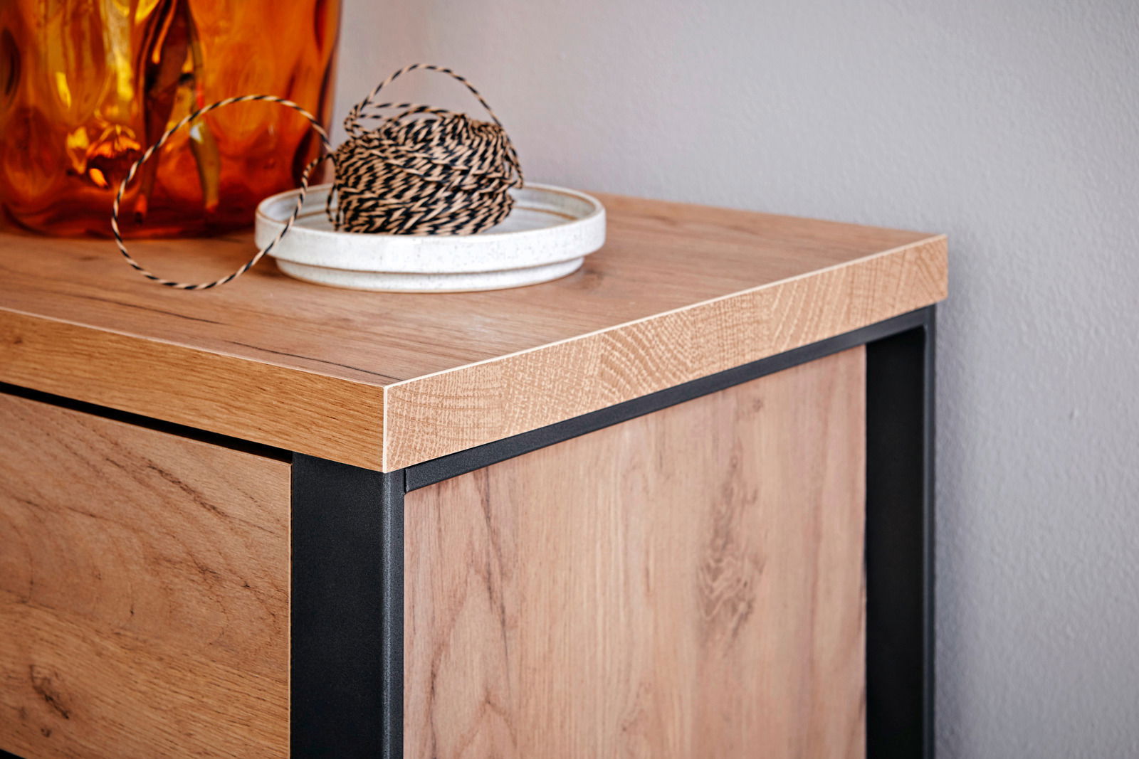 Nahaufnahme einer Ecke eines Eck-Schreibtisches aus Holz mit schwarzem Metallrahmen, dekoriert mit einer orangefarbenen Vase und einer Schale mit Schnur.