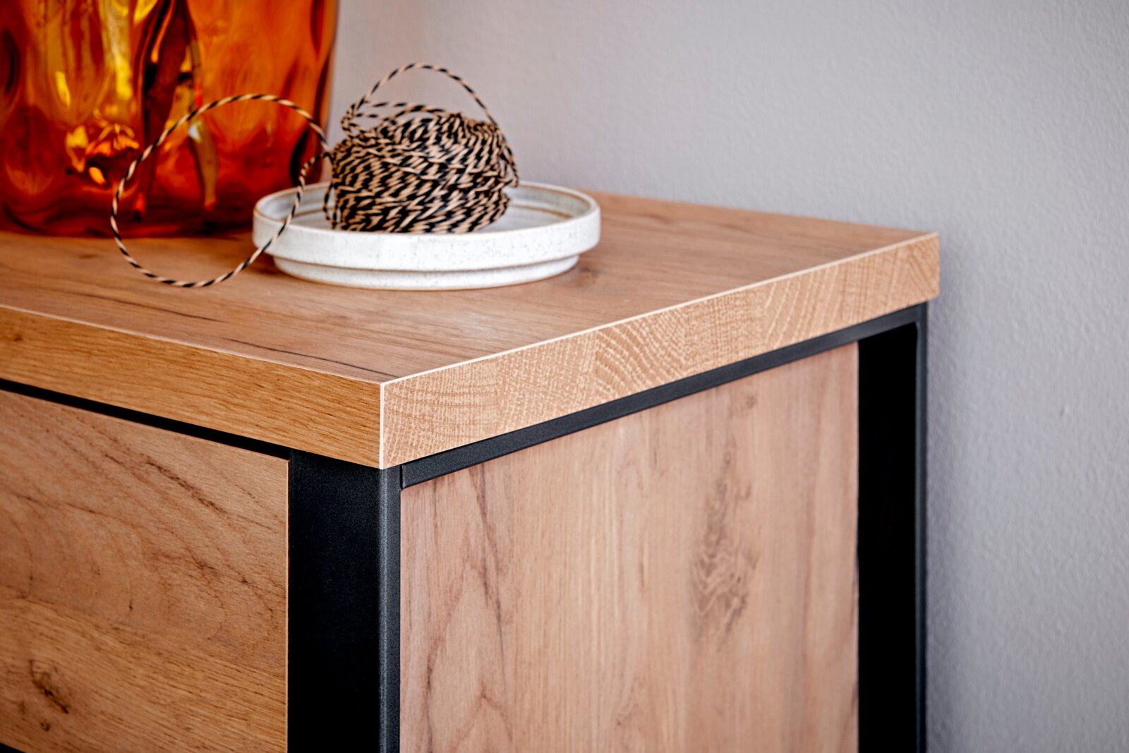 Close-up van een hoek van een houten hoekbureau met een zwart metalen frame, versierd met een oranje vaas en een schaal met touw.