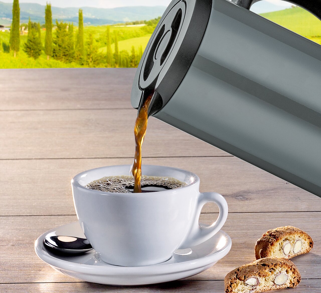 Graue Isolierkanne, die Kaffee in eine weiße Tasse auf einem Holztisch mit italienischer Landschaft im Hintergrund gießt, seitliche Perspektive.