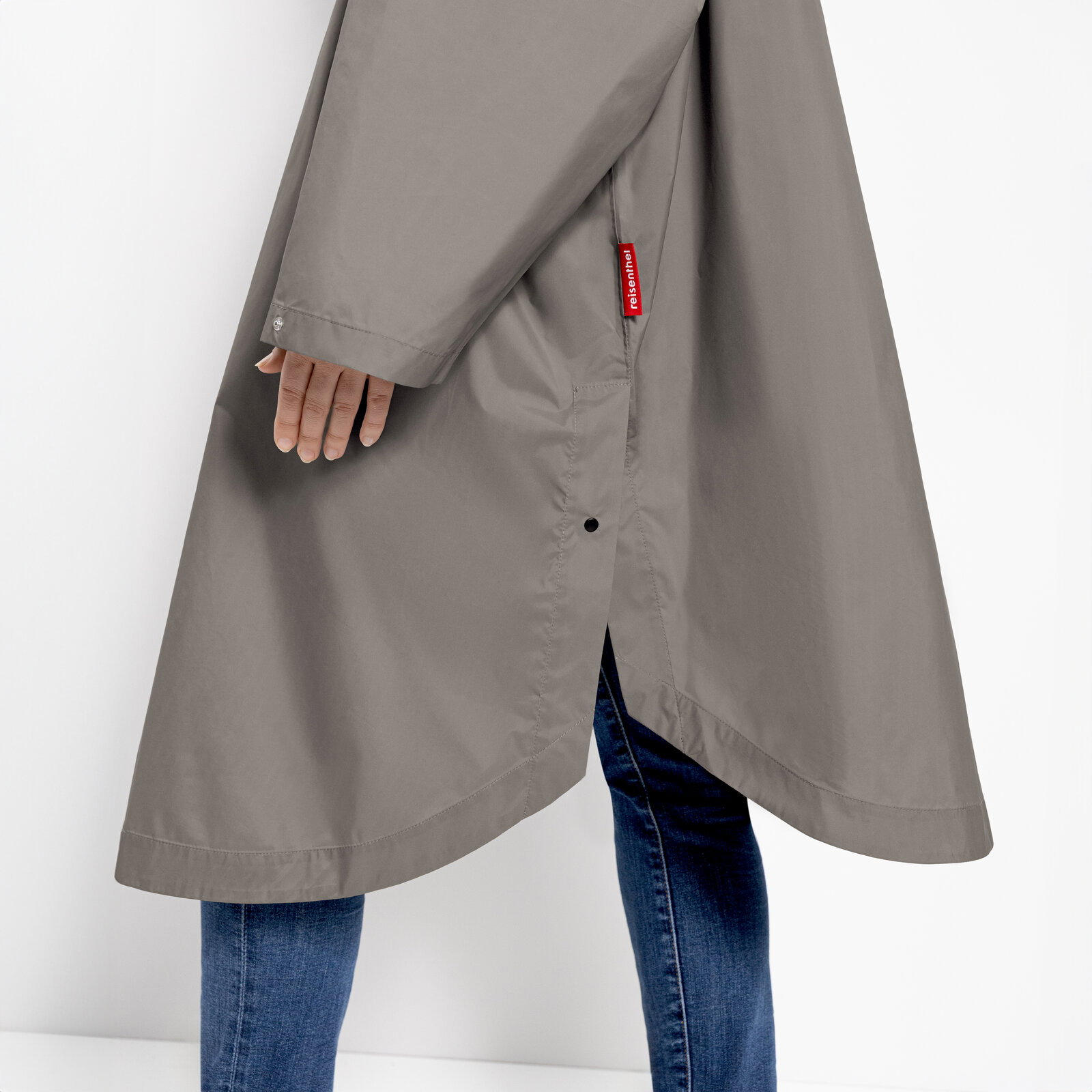 Seitliche Detailansicht eines grauen Regenmantels mit seitlichem Schlitz, Druckknopf und rotem reisenthel-Label, getragen über blauer Jeans