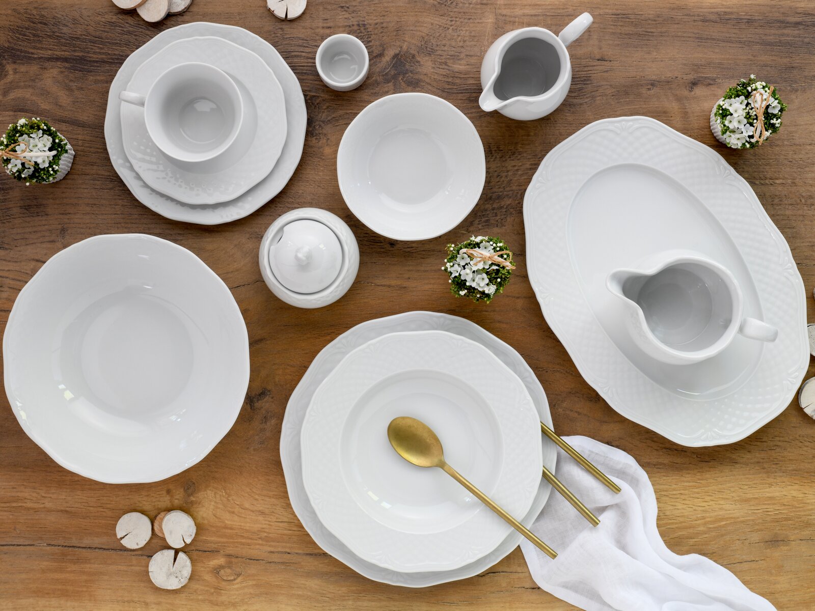 Draufsicht auf ein elegantes weißes Porzellan-Geschirrset mit Tellern, Schalen, Tassen, Zuckerdose und Milchkännchen auf rustikalem Holztisch, dekoriert mit kleinen Blumen und goldfarbenem Besteck.