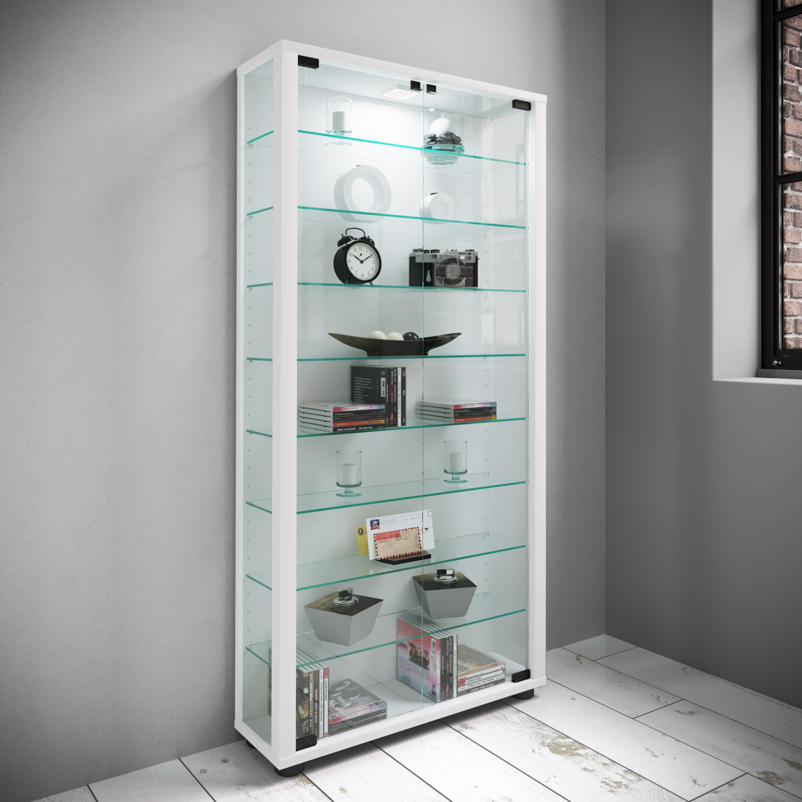 Weiße Glasvitrine mit LED-Beleuchtung und mehreren Glasböden, befüllt mit Deko und Büchern, in einem modernen Raum – Ansicht schräg von vorne links.