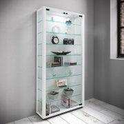 Weiße Glasvitrine mit LED-Beleuchtung und mehreren Glasböden, befüllt mit Deko und Büchern, in einem modernen Raum – Ansicht schräg von vorne links.