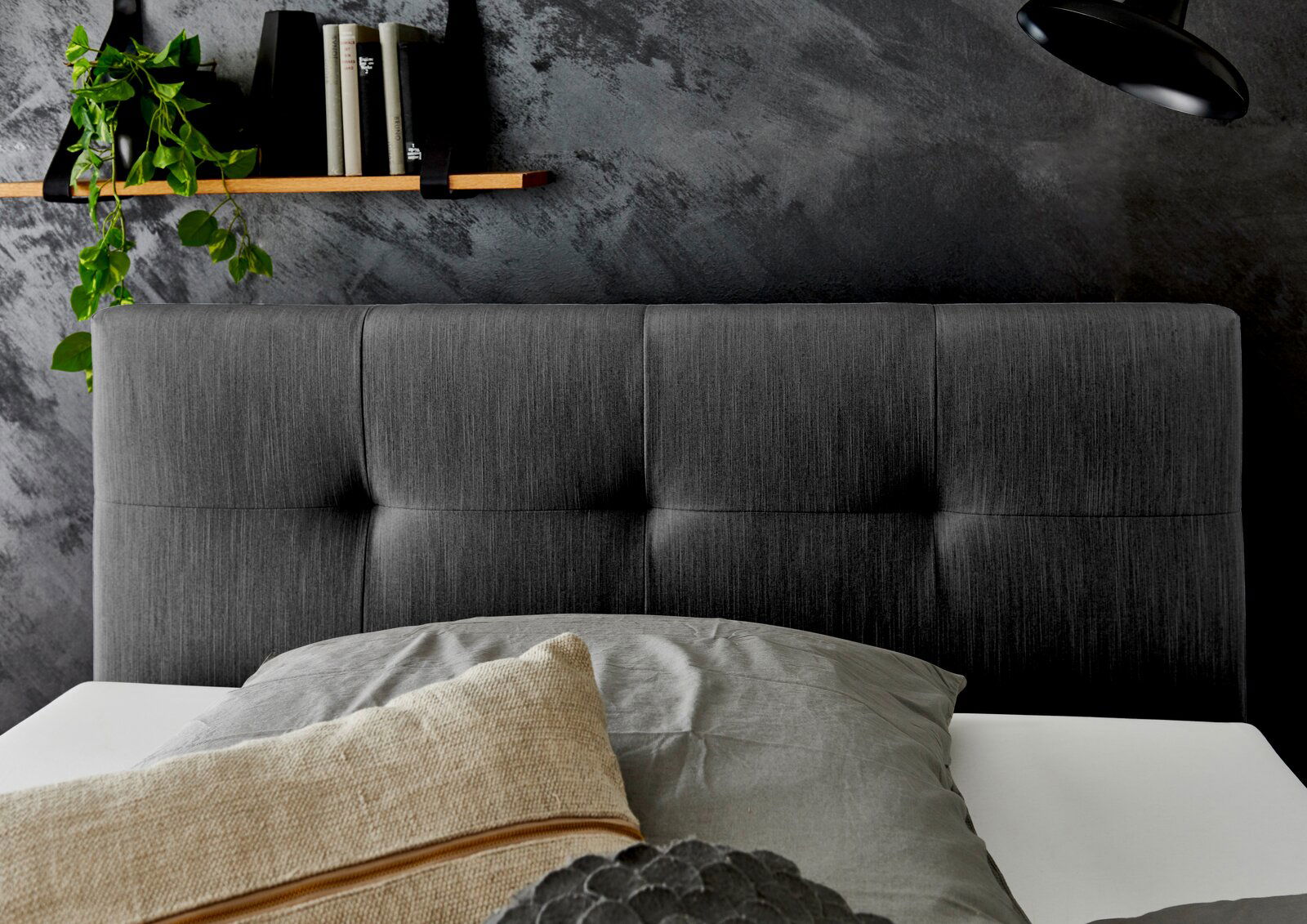 Boxspringbett mit grauem Kopfteil, dekoriert mit Kissen, im modernen Schlafzimmer mit dunkler Wand und Regal im Hintergrund, seitliche Perspektive