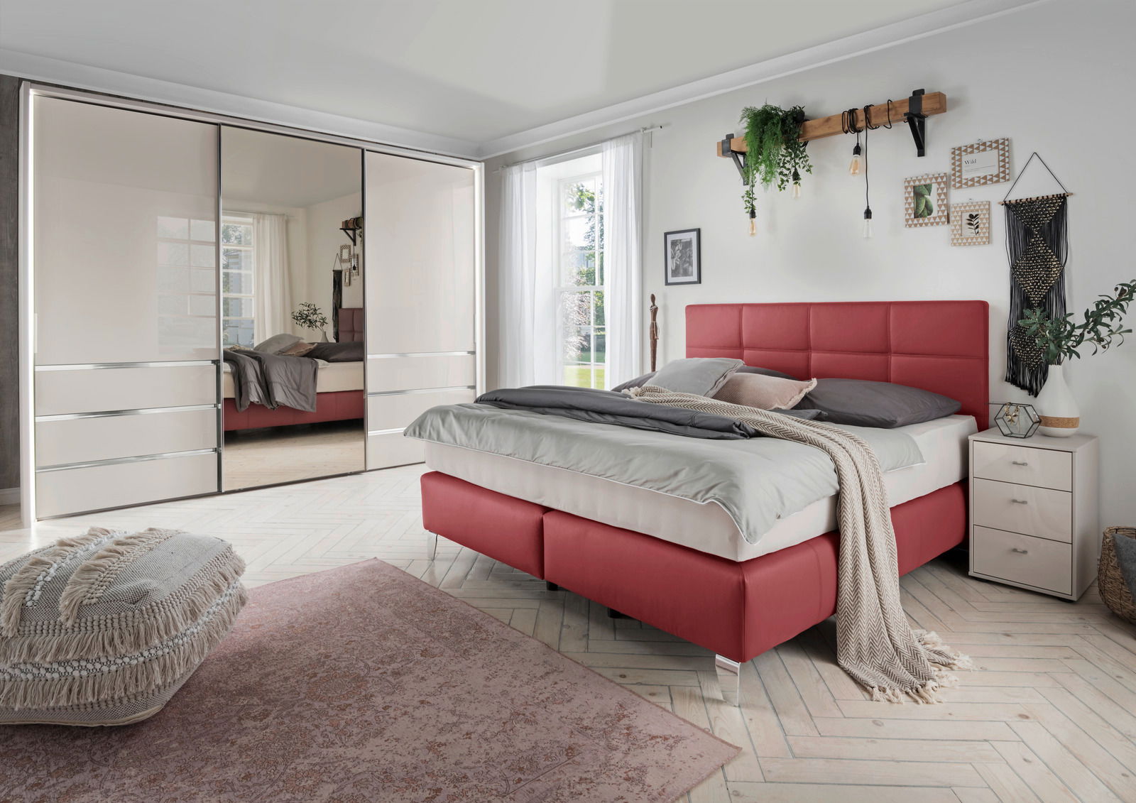 Schlafzimmer mit modernem Schwebetürenschrank in heller Farbe, Spiegel in der Mitte, rotem Polsterbett und dekorativen Elementen, seitliche Perspektive.