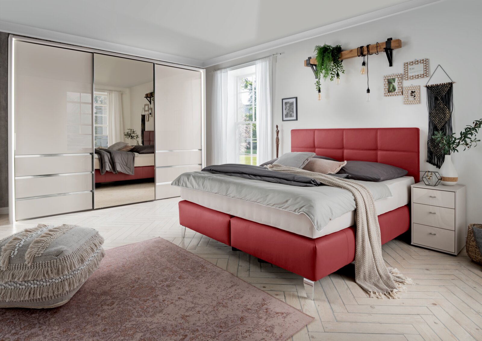Schlafzimmer mit modernem Schwebetürenschrank in heller Farbe, Spiegel in der Mitte, rotem Polsterbett und dekorativen Elementen, seitliche Perspektive.