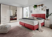 Schlafzimmer mit modernem Schwebetürenschrank in heller Farbe, Spiegel in der Mitte, rotem Polsterbett und dekorativen Elementen, seitliche Perspektive.