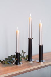 Drei schwarze, pulverbeschichtete Kerzenhalter in unterschiedlicher Höhe mit brennenden Kerzen, auf einem Holztisch mit Eukalyptuszweigen, seitliche Perspektive.