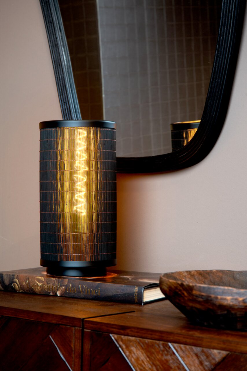 Moderne Tischlampe mit schwarzem, zylindrischem Schirm, auf einem Buch stehend, mit Spiegel im Hintergrund, seitliche Perspektive.
