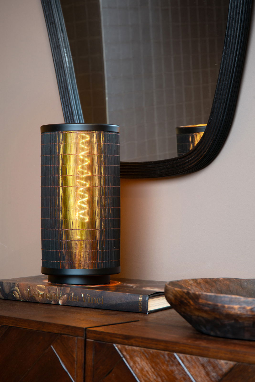 Moderne Tischlampe mit schwarzem, zylindrischem Schirm, auf einem Buch stehend, mit Spiegel im Hintergrund, seitliche Perspektive.