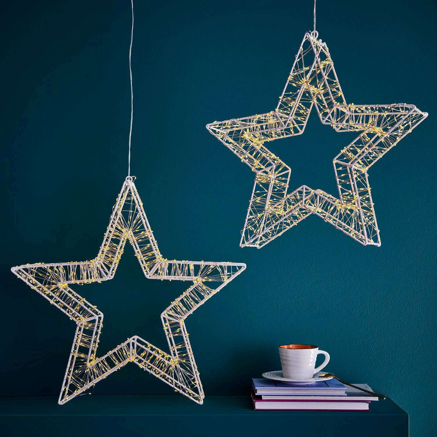 Zwei hängende LED-Sterne mit warmweißen Lichtern vor einer blauen Wand, seitliche Perspektive.