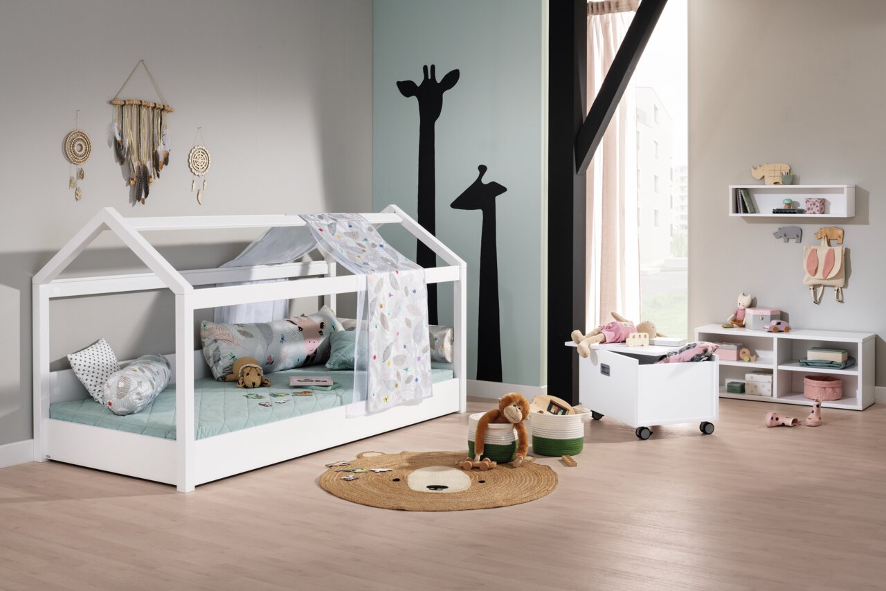 Een rolcontainer op wieltjes in een kinderkamer, gevuld met speelgoed, naast een wit huisbed en planken. Perspectief: zijaanzicht.