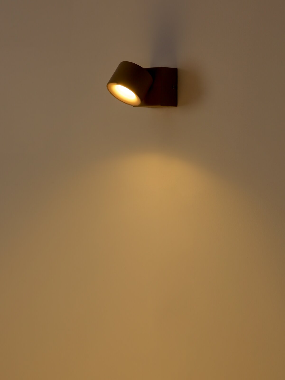 Schwarze, moderne Wandleuchte mit zylindrischem Spot, eingeschaltet und mit warmweißem Lichtkegel nach unten an einer hellen Wand, perspektivisch in leichter Seitenansicht von schräg vorn aufgenommen.