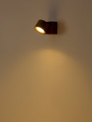 Schwarze, moderne Wandleuchte mit zylindrischem Spot, eingeschaltet und mit warmweißem Lichtkegel nach unten an einer hellen Wand, perspektivisch in leichter Seitenansicht von schräg vorn aufgenommen.
