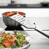 Grobreibe PRIMELINE aus Edelstahl, seitliche Perspektive, auf einer Arbeitsfläche mit einer Schüssel und einem Teller mit Salat.