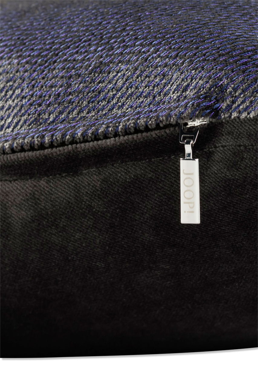 Nahaufnahme einer Zierkissenhülle mit feinem, strukturiertem Stoff in dunklen Tönen und einem silbernen Reißverschlussanhänger mit der Aufschrift 'JOOP!'.