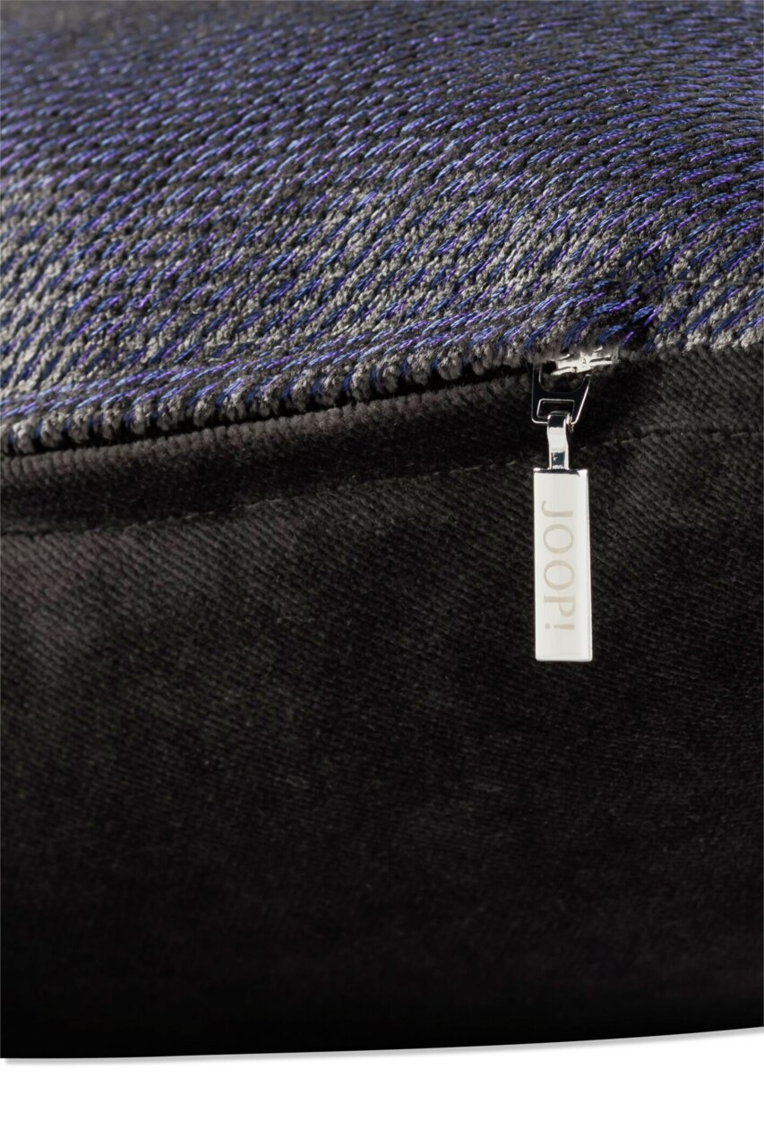 Nahaufnahme einer Zierkissenhülle mit feinem, strukturiertem Stoff in dunklen Tönen und einem silbernen Reißverschlussanhänger mit der Aufschrift 'JOOP!'.