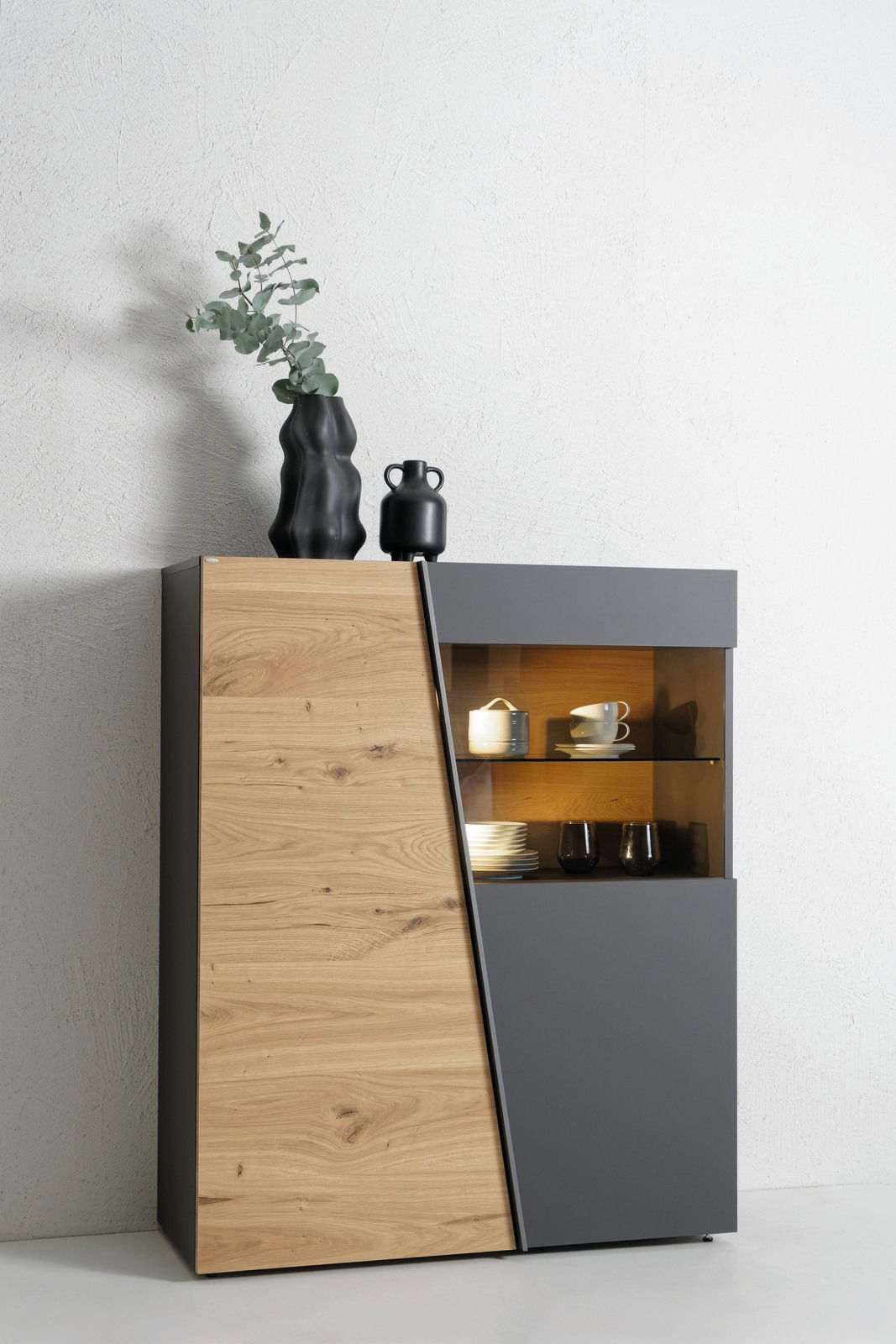 Modernes Highboard aus Holz und dunklem Material mit asymmetrischem Design, dekoriert mit schwarzen Vasen und Geschirr, seitliche Perspektive.