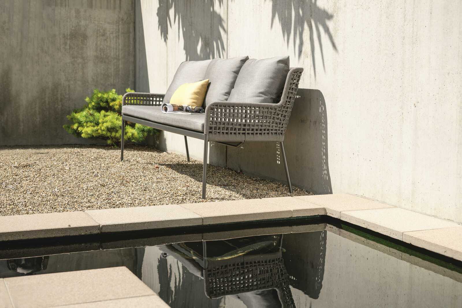 Stern Dining Bank GRETA Seitliche Perspektive auf eine stilvolle Dining Bank im Freien, mit grauen Kissen und einem gelben Akzentkissen, platziert auf einem Kiesboden neben einer Betonwand.