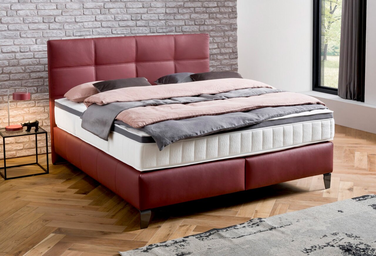 Boxspringbett in modernem Schlafzimmer, seitliche Perspektive, mit rotem Kopfteil und Bettgestell, graue und rosa Bettwäsche, Ziegelwand im Hintergrund.
