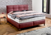 Boxspringbett in modernem Schlafzimmer, seitliche Perspektive, mit rotem Kopfteil und Bettgestell, graue und rosa Bettwäsche, Ziegelwand im Hintergrund.