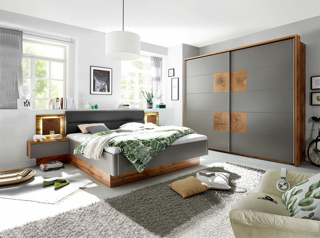 Moderne Schlafzimmeransicht mit Bettanlage in Holzoptik und grauem Kopfteil, flankiert von Nachttischen mit Beleuchtung, im Vordergrund ein gemütlicher Teppich und im Hintergrund ein großer Kleiderschrank mit Holzdetails.