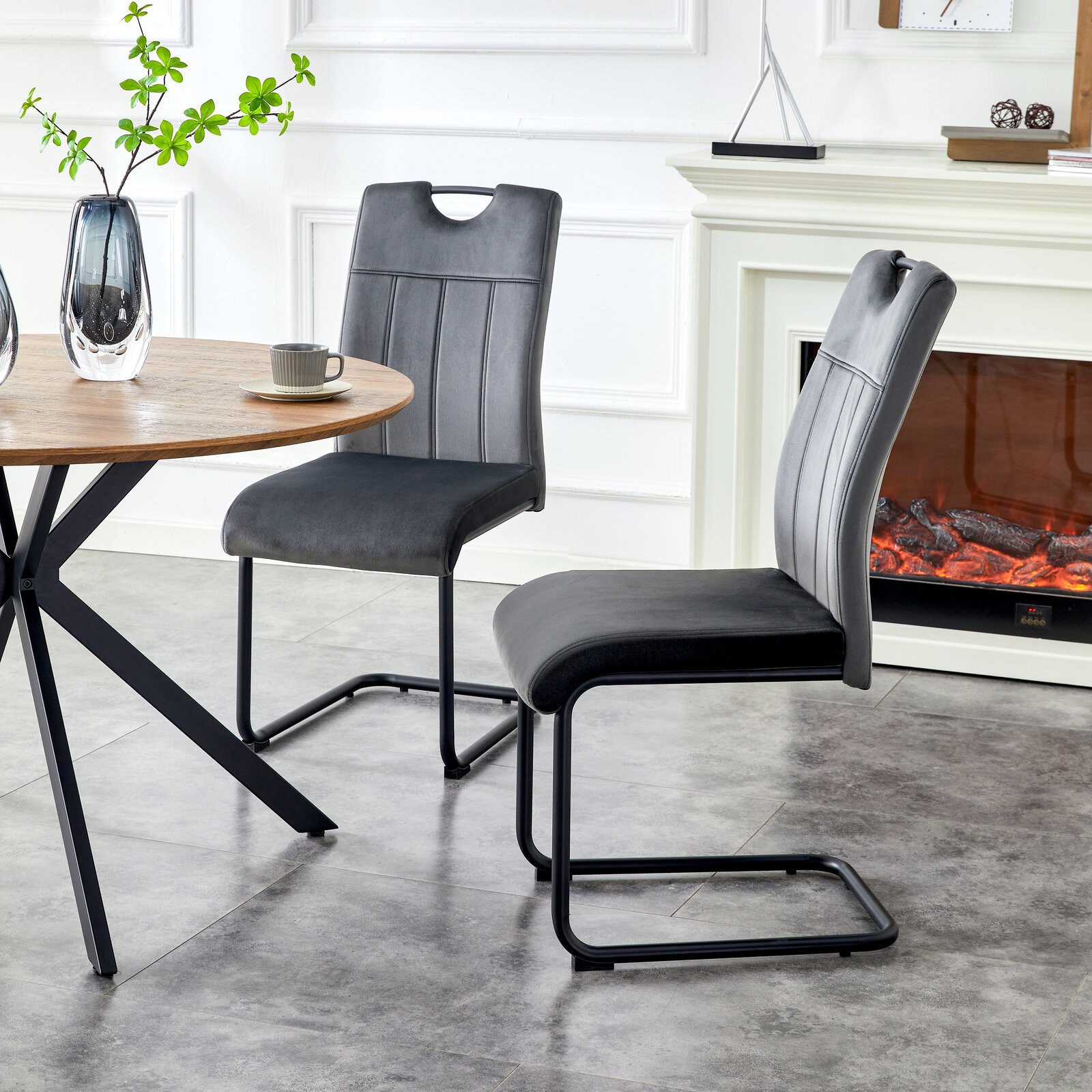 Zwei moderne Freischwinger-Esszimmerstühle in Dunkelgrau mit gepolsterter Sitzfläche und Rückenlehne, schwarzem Metallgestell und Ziernähten, seitliche Perspektive im Wohn-/Essbereich neben rundem Holztisch und Kamin.