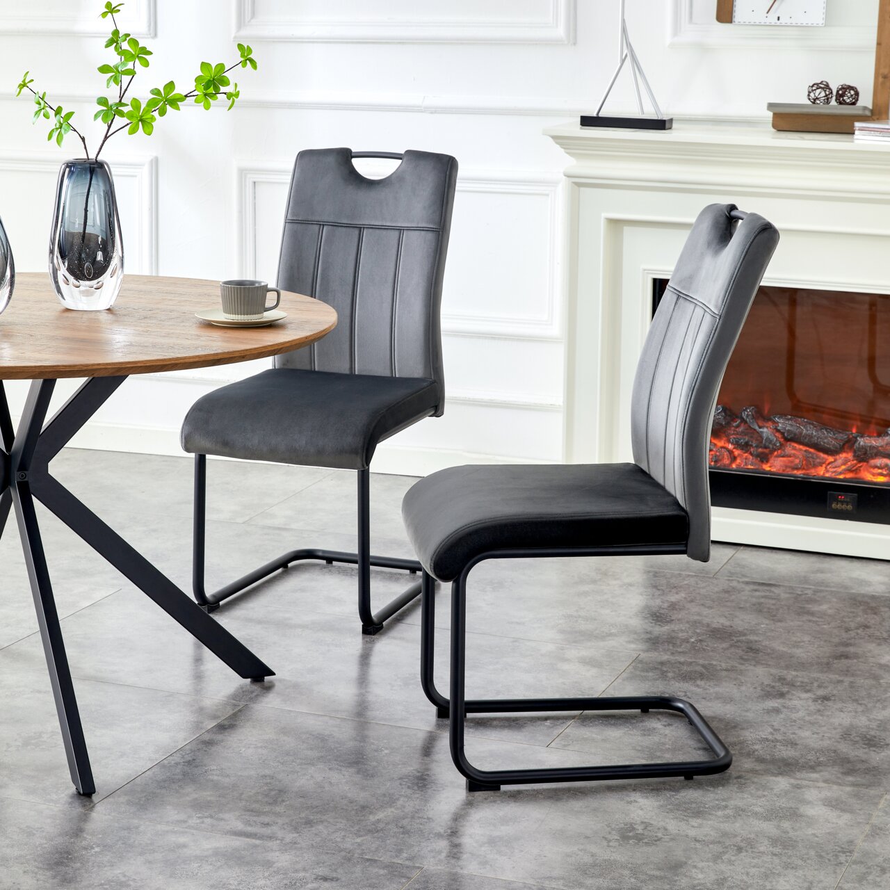 MID.YOU Schwingstuhl 2er-Set LISA Zwei moderne Freischwinger-Esszimmerstühle in Dunkelgrau mit gepolsterter Sitzfläche und Rückenlehne, schwarzem Metallgestell und Ziernähten, seitliche Perspektive im Wohn-/Essbereich neben rundem Holztisch und Kamin.