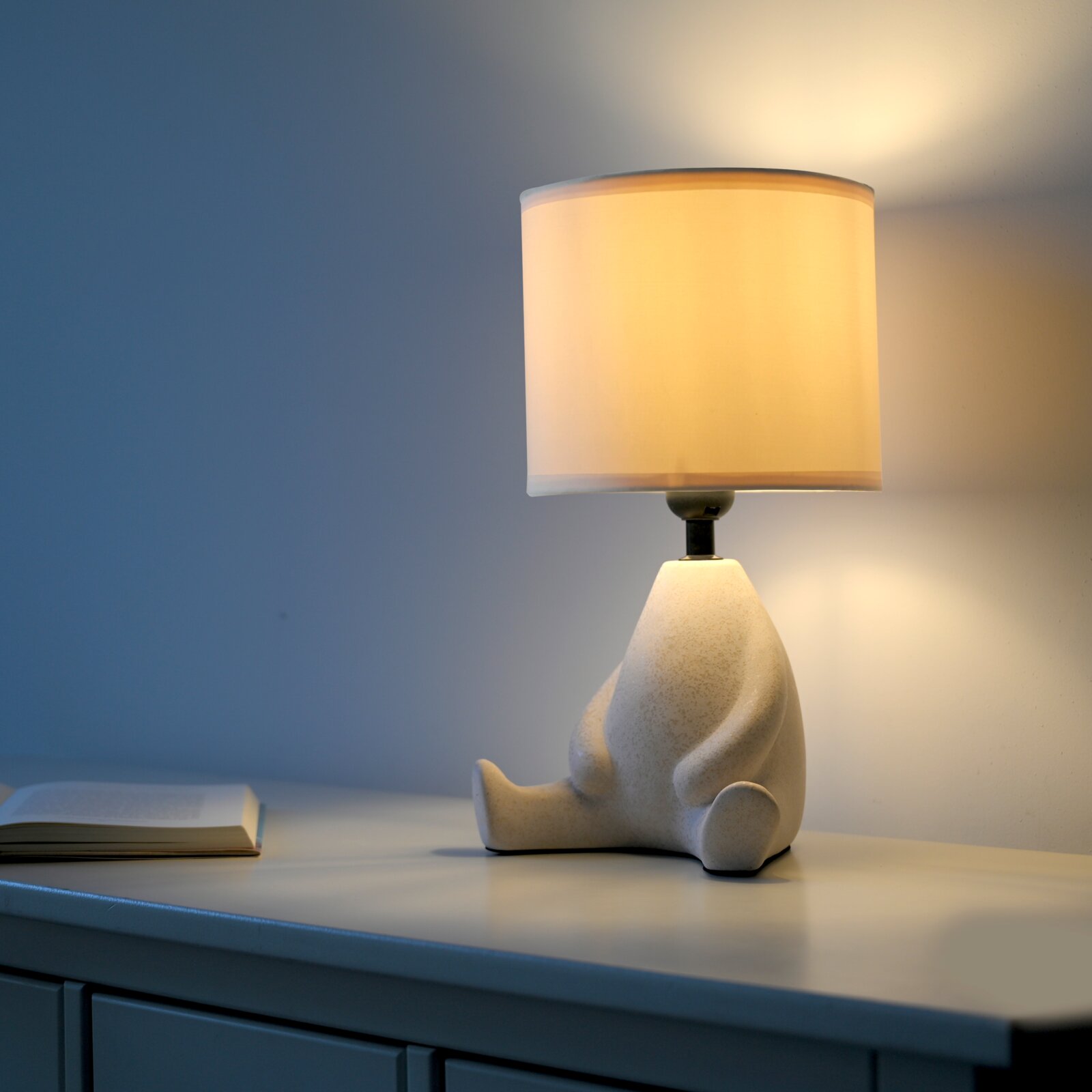 Leuchtende Tischlampe mit weißem zylindrischem Stoffschirm und skulpturalem Keramikfuß auf Sideboard, seitliche Ansicht aus leicht erhöhter Perspektive