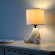 Leuchtende Tischlampe mit weißem zylindrischem Stoffschirm und skulpturalem Keramikfuß auf Sideboard, seitliche Ansicht aus leicht erhöhter Perspektive