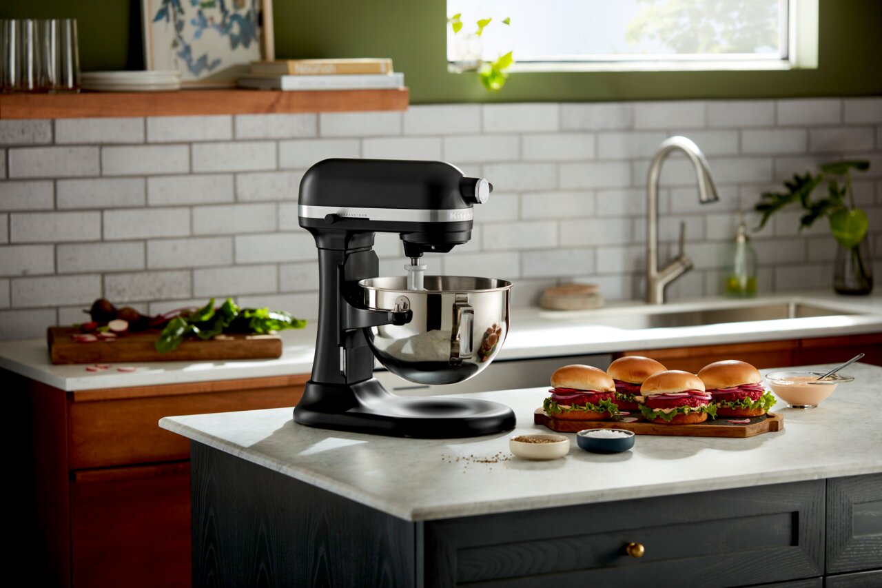 KitchenAid Keukenmachine HEAVY DUTY 5KSM55SXXEBM Zwarte keukenmachine Heavy Duty 5,2 l op een aanrechtblad, zijaanzicht, met een mengkom en een deeghaak, omringd door verse ingrediënten en broodjes.