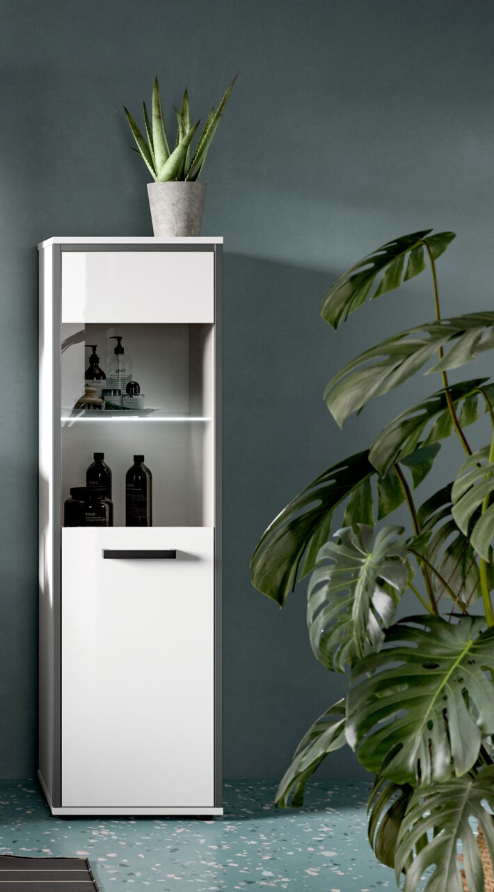 Schmaler, hoher Badezimmerschrank in Weiß mit Glastür und LED-Beleuchtung, unten geschlossene Tür mit schwarzem Griff, mit Pflanze obenauf; frontale Ansicht leicht von links vor grauer Wand.