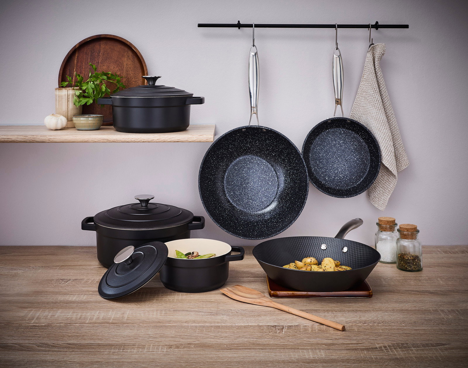 Küchenszene mit mehreren Kochutensilien, darunter eine 20cm Alu-Pfanne mit Antihaftbeschichtung, die an einer Stange hängt. Perspektive von vorne.