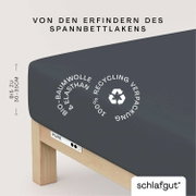 Spannbettlaken aus Bio-Baumwolle und Elasthan in dunkler Farbe, auf einem Bett mit Holzrahmen. Verpackung aus 100% Recyclingmaterial. Perspektive von der Seite.