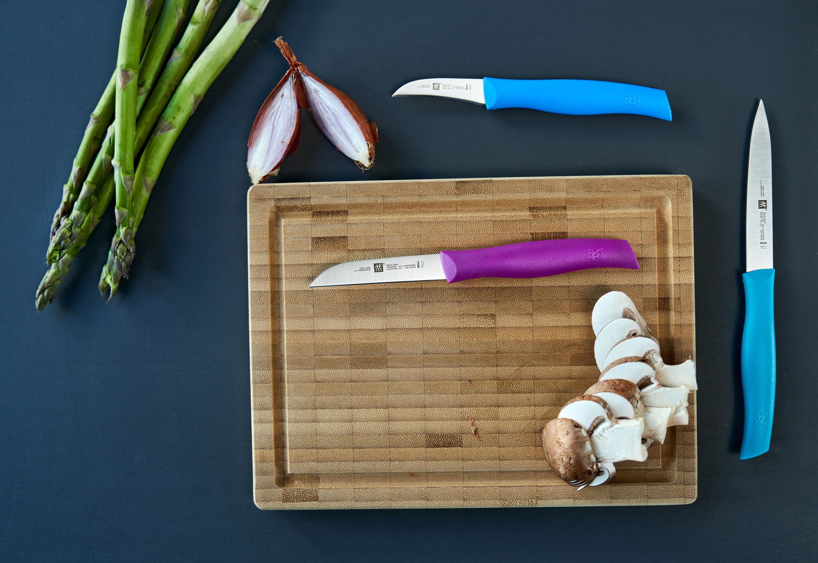 Draufsicht auf ein dreiteiliges Messerset mit bunten Griffen auf einem Schneidebrett, umgeben von Spargel, Zwiebelhälften und geschnittenen Pilzen.